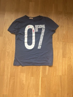 2012 marinblå Haikuretröja - Säljer en blå t-shirt med stor vit 07-print och textdetaljer framtill. T-shirten har korta ärmar och en klassisk passform. Perfekt för dig som gillar grafiska tryck och enkel stil.