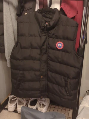 Svart dunväst från Canada Goose - Knapparna där nere hålls inte stängda när man knäpper den därav priset nygg svart dunväst från Canada Goose med klassisk logga på bröstet. Västen har hög krage, två fickor framtill och stängs med dragkedja och knappar. Perfekt för lager-på-lager och håller dig varm under kyliga dagar.