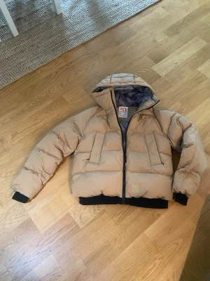 Beige pufferjacka med huva - Snygg beige pufferjacka med huva och svarta muddar. Jackan har två stora fickor framtill med dragkedja och en quiltad insida i grått. Perfekt för kalla vinterdagar och riktigt trendig look. Nypris 2500:-  säljes för endast 1500:-!