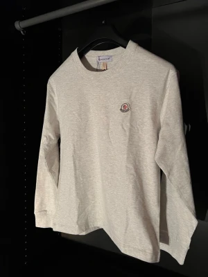 Ljusgrå långärmad topp från Moncler - Unik Ljusgrå långärmad topp från Moncler med rund halsringning och diskret Moncler-logga på bröstet. Tillverkad i mjuk bomull som känns skön mot huden. Enkel och stilren design som passar till många olika outfits.