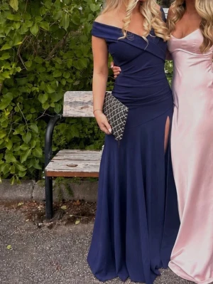 Marinblå långklänning offshoulder - Elegant marinblå långklänning med offshoulder-skärning och figurnära passform. Klänningen har en snygg slits på ena sidan och omlott-detaljer vid bysten. Perfekt för bal eller festliga tillfällen.