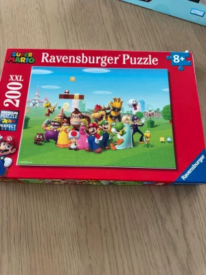 Super Mario pussel Ravensburger 200 bitar - Super Mario pussel från Ravensburger med 200 XXL-bitar. Motivet visar flera klassiska karaktärer från Super Mario-världen på en grön äng med slott i bakgrunden. Lådan är röd och blå med tydliga Super Mario-detaljer. Passar för åldrar 8+.