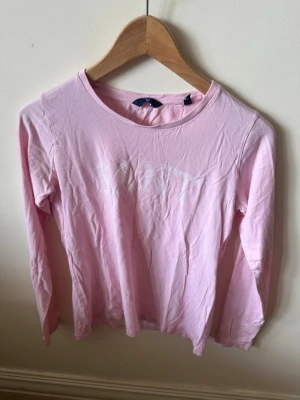 Ljusrosa långärmad topp från GANT - Ljusrosa långärmad topp från GANT med rund halsringning och vit logga tryckt framtill. Tillverkad i mjuk bomull som känns skön mot huden. Perfekt att styla med jeans eller kjol för en enkel och snygg look.