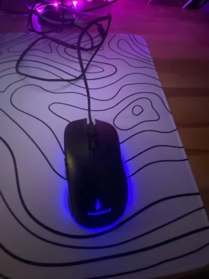 Svart gamingmus med flera olika LED-belysningar som tex blå, rosa, gul, grön och röd,  - En svart gamingmus med sladd och blå LED-belysning undertill. Musen har en slimmad, ergonomisk form och två knappar på ovansidan. Passar perfekt för gaming eller arbete vid datorn. Materialet är plast och designen är stilren.