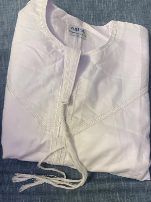 Vit traditionell kaftan med brodyr - Säljer en vit traditionell kaftan med dekorativa broderier och snörning framtill. Tillverkad i ett lätt och luftigt tyg, perfekt för dig som gillar stilrena och bekväma plagg. Passar till många olika tillfällen och har en klassisk design.