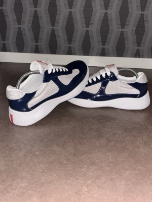 Vita och marinblå sneakers - Snygga sneakers i vitt och marinblått med meshpaneler och lackdetaljer. Skorna har vita snören, tjock vit sula och en röd detalj på hälen. Perfekta för dig som gillar en sportig och fräsch stil.