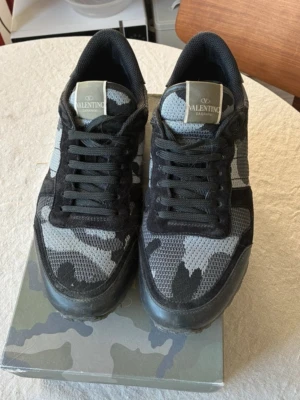 Valentino camo sneakers i svart/grå - Snygga sneakers från Valentino med svart och grått camomönster. Skorna har mesh- och mockadetaljer, svarta skosnören och en robust sula. Perfekta för dig som vill ha en unik och trendig look.