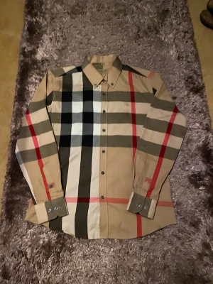 Burberry skjorta - Säljer en beige Burberry-skjorta i storlek M. Helt ny och i perfekt skick. En elegant skjorta som passar lika bra till dejter, middagar, fester eller andra tillfällen där du vill klä upp dig. Lätt att matcha och ger en riktigt stilren look.