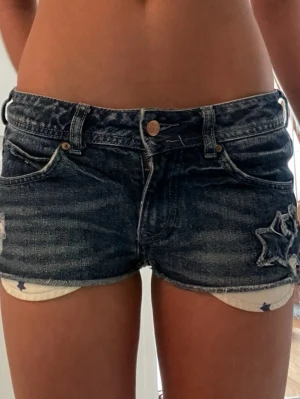 Mörkblå låga jeansshorts med stjärndetaljer - Säljer ett par mörkblå jeansshorts med slitningar och stjärnformade detaljer på ena benet. Shortsen har klassisk femficksdesign, knappgylf och råa kanter. Fickorna har ett vit tyg med blåa stjärnor.