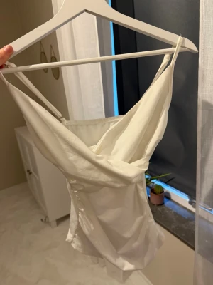 Vit satinliknande topp med smala band - Snygg vit topp med tunna axelband och draperad urringning. Materialet har en satinliknande finish som ger ett lyxigt intryck. Perfekt att styla med jeans eller kjol för en trendig look.