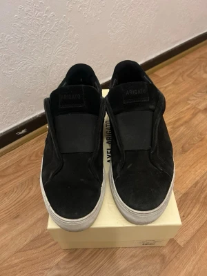 Svarta slip-on sneakers Axel Arigato - Stilrena svarta slip-on sneakers från Axel Arigato med ovandel i mocka och bred resår över vristen. Skorna har vit sula och minimalistisk design utan snörning. Perfekta för en clean och modern look lådan följer med jätte fin skick pris kan diskuteras vid en snabb och smidig affär .