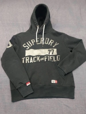 Mörkgrå Superdry hoodie med tryck - Snygg mörkgrå hoodie från Superdry med stort vitt tryck 'Track and Field 77' på bröstet. Klassisk huva med vita snören och känguruficka framtill. Ribbade muddar och detaljerade Superdry-märken på ärm och nederkant.