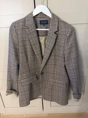 Rutig kavaj från Lexington - Snygg rutig kavaj från Lexington i beige, brunt och vitt. Klassisk modell med två framfickor, en knapp och slag. Fodrad insida och uppvikta ärmar för en avslappnad look. Perfekt för dig som gillar tidlös stil och vill sticka ut lite extra.