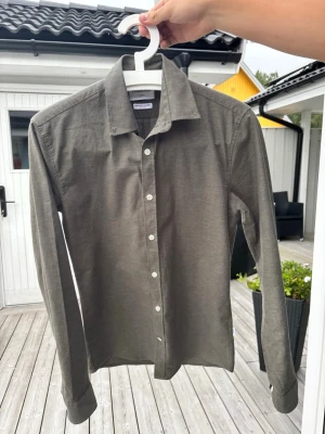 Lindbergh skjorta strl S slim fit - Stilren olivgrön skjorta i bomull med klassisk krage och vita knappar framtill. Skjortan har lång ärm och slim fit som funkar till många olika looks. Perfekt för dig som gillar en enkel men snygg stil.