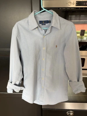 Ljusblå skjorta från Ralph Lauren - Klassisk ljusblå skjorta från Ralph Lauren med button-down krage och lång ärm. Skjortan har den ikoniska broderade loggan på bröstet och vita knappar framtill. Tillverkad i mjukt bomullstyg och har en avslappnad passform.