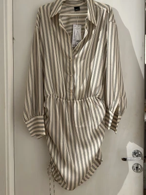 Randig skjortklänning från Gina Tricot - Säljer en beige och vit randig skjortklänning från Gina Tricot. Klänningen har långa ärmar, krage, knappar framtill och snörning i midjan. Materialet är mjukt och känns som polyester. Perfekt för dig som gillar en avslappnad men trendig stil. Passar Stl M oxå.