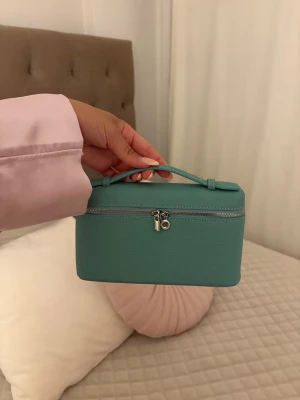 Turkos boxig handväska med handtag - Snygg turkos handväska i boxig modell med ett smidigt handtag upptill. Väskan har silverfärgad dragkedja och är gjord i ett slätt, strukturerat material som liknar konstläder. Perfekt för dig som gillar stilrena accessoarer med en poppig färg.