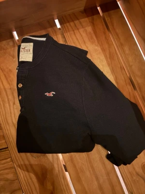 Svart långärmad tröja Hollister - Svart långärmad tröja från Hollister med knappslå och diskret logga på bröstet. Tröjan har ribbade muddar och är tillverkad i ett mjukt, strukturerat material som ger en avslappnad look.