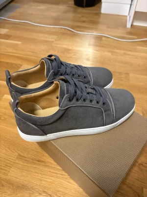 Gråa sneakers från Christian Louboutin - Snygga gråa sneakers i mocka från Christian Louboutin med klassisk röd sula och vita kanter. Skorna har rund tå, matchande grå skosnören och diskreta sömdetaljer. Perfekta för dig som vill ha en stilren och exklusiv look, och har använt dem 2 gånger