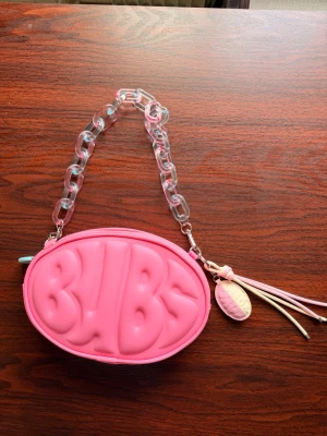 Rosa oval handväska med kedjehandtag - Cool rosa handväska i oval form med upphöjd BUBS-text på framsidan. Väskan har ett transparent rosa kedjehandtag och dragkedja med blått BUBS-märke. Dekorativa tofsar och godisberlock i pastellfärger hänger på sidan. Materialet är konstläder.