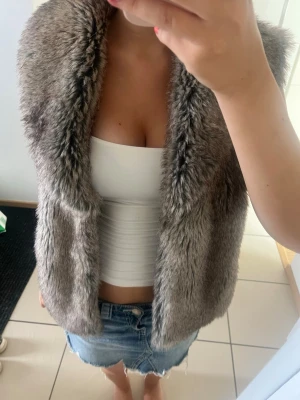 Grå pälsväst från CNY Outerwear - Snygg grå pälsväst från CNY Outerwear i storlek M. Västen har en mjuk och fluffig fuskpäls med öppen front och slät insida. Perfekt att slänga över en topp för en trendig look. Passar dig som vill ha något extra till din outfit.