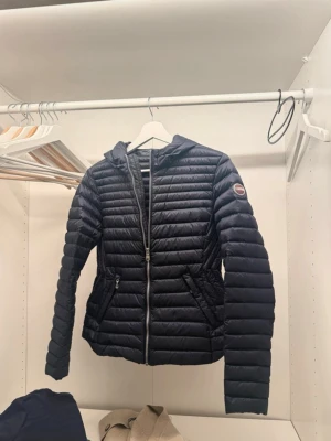 Marinblå Colmar  dunjacka med huva - Snygg  marinblå dunjacka med huva och quiltad design. Jackan har dragkedja framtill, två sidofickor med dragkedja och en patch på ärmen. Perfekt för kalla vinter höst dagar och ger en stilren look. Köpt för runt 3000kr