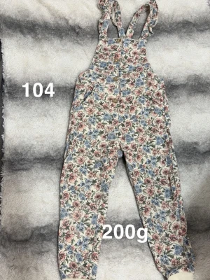 Blommig byxdress med volangband - Byxdress med blommigt mönster i rosa, blått och grönt. Axelbanden har volangdetaljer och byxdressen har fickor på sidorna. Materialet ser ut att vara mjukt och bekvämt, perfekt för en lekfull och trendig look.🌸 Använd en gång. Storlek 104 