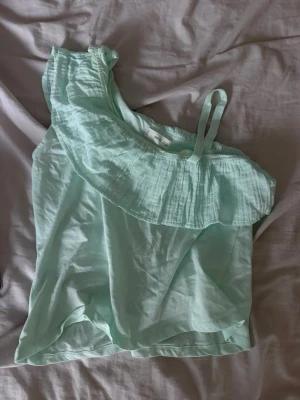 Mintgrön oneshoulder topp med volang - Superfin mintgrön topp med oneshoulder-design och volangdetalj längs halsringningen. Toppen är ärmlös med ett axelband och har en luftig känsla, perfekt för varma dagar.