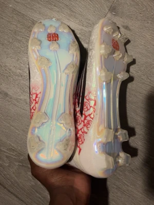 Vita fotbollsskor med rosdetaljer - Säljer ett par vita fotbollsskor med röda rosdetaljer längs ovandelen. Skorna har en låg profil och mönstrad sula med fasta dobbar i genomskinligt material. Perfekta för dig som vill sticka ut på planen.