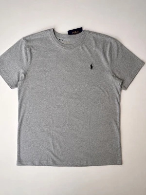 Grå t-shirt från Polo Ralph Lauren LARGE - Klassisk grå t-shirt från Polo Ralph Lauren med rund halsringning och diskret svart logobrodyr på bröstet. Tillverkad i mjuk bomull för en bekväm känsla. Aldrig använd med taggar