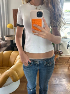 Vit croppad t-shirt med svarta detaljer - Säljer en vit croppad t-shirt med svarta ribbade kanter vid ärmsluten. T-shirten har en tight passform och rund halsringning. Perfekt till jeans eller kjol för en enkel och snygg look. Missfärgning på bild 3.