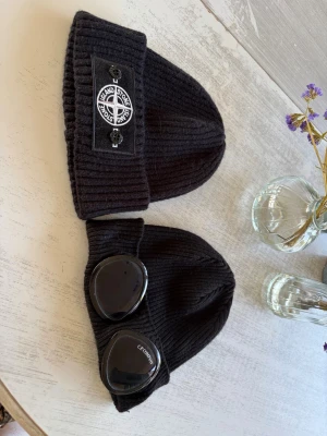 Två svarta ribbade mössor - Säljer två svarta ribbade mössor (använt mycket få gånger). Ena har en patch med Stone Island-inspirerad logga framtill, den andra har två stora svarta glasögon-liknande detaljer från C.P. Company. Båda är stickade och har uppvikt kant. Perfekta för dig som gillar streetwear. 