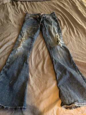Blå slitna bootcut jeans - Säljer ett par blå bootcut jeans med slitningar och hål på båda benen. Lågmidjade storlek xs. På vänster ben är hålet lite större för de har gått upp lite annars inga defekter. 