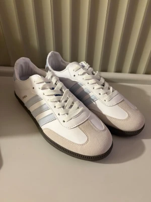 Vita Adidas Samba sneakers - Säljer ett par klassiska Adidas Samba sneakers i vitt med ljusblå detaljer och de ikoniska tre ränderna på sidan. Skorna har en låg silhuett, snörning och svart yttersula. Materialet är en mix av läder och mocka för en stilren look.