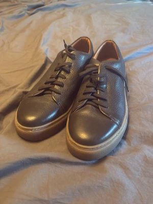 Svarta sneakers från Doppelgänger - Stilrena svarta sneakers från Doppelgänger i slätt och lätt texturerat läder. Skorna har svarta snören, bruna innersulor med guldfärgad logga och en kontrasterande beige sula. Perfekta för dig som vill ha en clean och klassisk look.