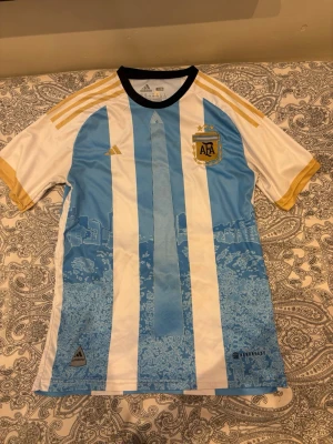 Argentina Messi matchtröja Adidas - Säljer en officiell Argentina fotbollströja från Adidas med Messi och nummer 10 på ryggen. Tröjan har klassiska blå och vita ränder, gulddetaljer på axlar och ärmslut samt AFA-emblem på bröstet. Tillverkad i lätt och ventilerande material, perfekt för match eller träning.