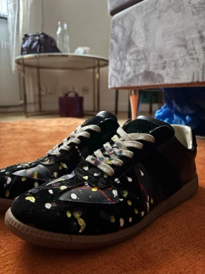 Maison Margiela svart sneakers med färgstänk - Unika svarta sneakers från Maison Margiela med färgstänk i vitt, gult, rött och blått. Skorna har en klassisk låg siluett, vita snören och detaljer i mocka och läder. Perfekta för dig som vill sticka ut med din stil.