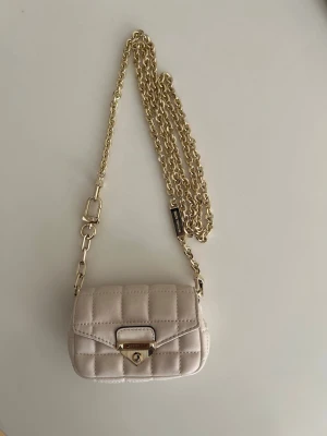 Beige quiltad mini handväska Michael Kors - Snygg beige quiltad mini handväska från Michael Kors med guldfärgad kedjerem och elegant låsdetalj framtill. Väskan har en kompakt form och stilren design som passar perfekt till trendiga outfits.