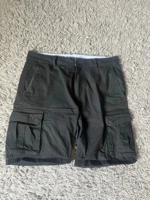 Svarta cargoshorts från Dressmann - Säljer ett par svarta cargoshorts från Dressmann med klassiska stora sidofickor och knappstängning. Shortsen har en avslappnad passform och är tillverkade i ett slitstarkt bomullsmaterial. Perfekta för en chill och bekväm stil.