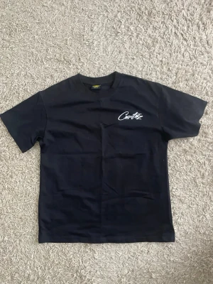 Svart cortiez t-shirt med vit text - En svart t-shirt med korta ärmar och vit text på bröstet. T-shirten har en klassisk passform och rund halsringning. Materialet känns som bomull och trycket ger en clean streetwear-vibe.