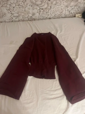 Vinröd stickad tröja med vida ärmar - Mysig vinröd stickad tröja med bred ribbad midja och extra vida ärmar. Tröjan har en avslappnad passform och rund halsringning. Perfekt för dig som gillar trendiga silhuetter och vill ha något bekvämt men ändå snyggt.