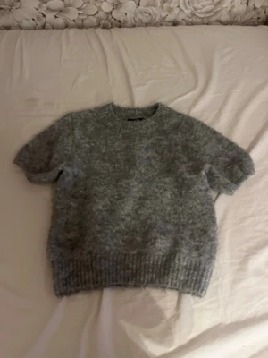 Grå stickad t-shirt - Mysig grå stickad t-shirt med korta ärmar och rund hals. Tröjan har en fluffig struktur och ribbstickade muddar vid ärmslut och nederkant. Perfekt för lager på lager och en avslappnad stil.