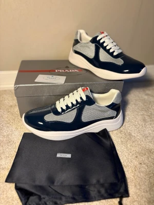 Sneakers i mörkblå, grått och vitt - Snygga  sneakers i mörkblå lack och vitt med meshdetaljer och vita skosnören. Skorna har en chunky vit sula och röda logodetaljer på plösen och sulan. Perfekta för dig som vill ha en exklusiv och modern look.