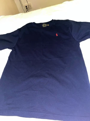 Mörkblå t-shirt från Polo Ralph Lauren - Säljer en mörkblå t-shirt från Polo Ralph Lauren med klassisk passform och rund hals. T-shirten har en liten röd broderad logga på bröstet och är tillverkad i mjuk bomull. Perfekt för en stilren och avslappnad look.