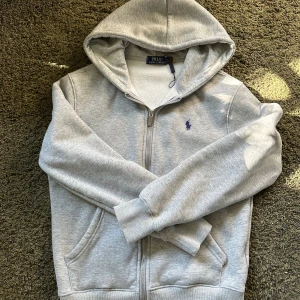 Grå hoodie från Polo Ralph Lauren - Snygg grå hoodie från Polo Ralph Lauren med dragkedja och huva. Tröjan har en broderad logga i mörkblått på bröstet, två fickor fram och mjukt innerfoder. Perfekt för chill dagar eller när du vill ha en clean look. Säljer pga att den inte passar på mig! .