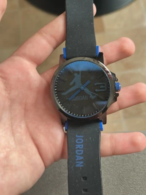 Svart Jordan klocka med blå  - Stilren svart klocka med blå detaljer och Jordan-logga på urtavlan. Klockan har ett svart armband i syntetmaterial med blå text och detaljer. Urtavlan är rund och har en sportig look med tydliga markeringar och blå krona. Helt ny inte använd säljer på grund behöver pengarna jätte bra 