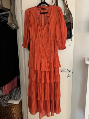 Maje klänning - Snygg långklänning i en stark orange färg med flera volanglager och v-ringning. Klänningen har trekvartsärm med lätt puff och smockdetaljer i midjan. Perfekt för sommarens alla tillfällen. Nypris 3500