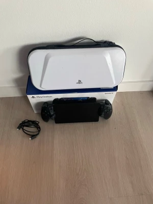 PlayStation Portal handhållen konsol - Säljer en PlayStation Portal handhållen spelkonsol från Sony. Konsolen är svart med integrerade kontroller på sidorna och har en stor skärm i mitten. Medföljer gör en vit skyddsväska och laddkabel. Perfekt för gaming på språng.