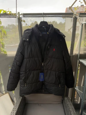  Polo Ralph Lauren vinter jacka - Snygg svart dunjacka från Polo Ralph Lauren med huva och dragkedja framtill. Jackan har två framfickor med dragkedja och den klassiska röda loggan på bröstet. Perfekt för kalla vinterdagar och riktigt stilren look.