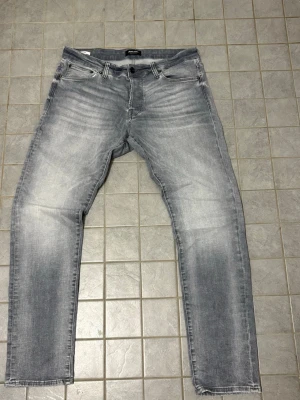 Grå slim fit jeans Slim/Glenn - Snygga grå slim fit jeans med slitningar och tvättade detaljer. Modellen heter Slim/Glenn och har klassisk femficksdesign, knappgylf och smal passform. Perfekta för en trendig och avslappnad stil.
